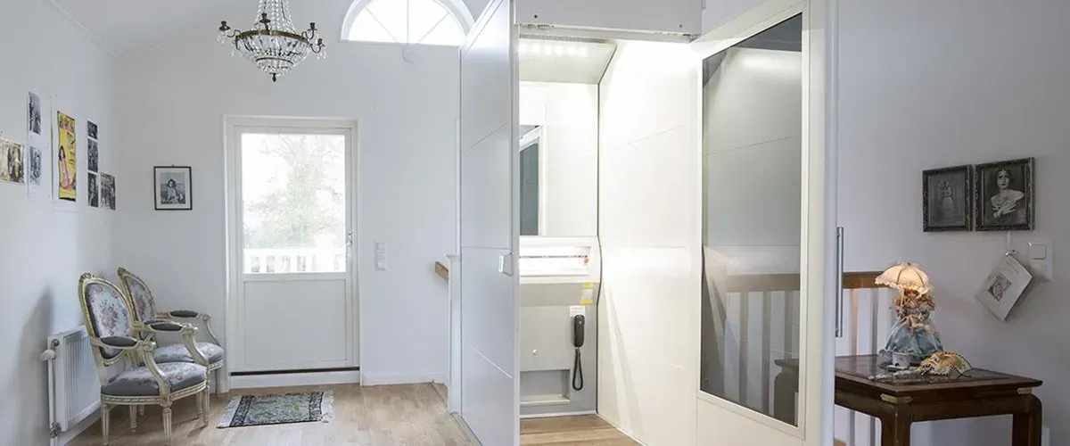 Un couloir blanc lumineux avec un lustre, des chaises et une porte entrouverte donnant sur une salle de bains.