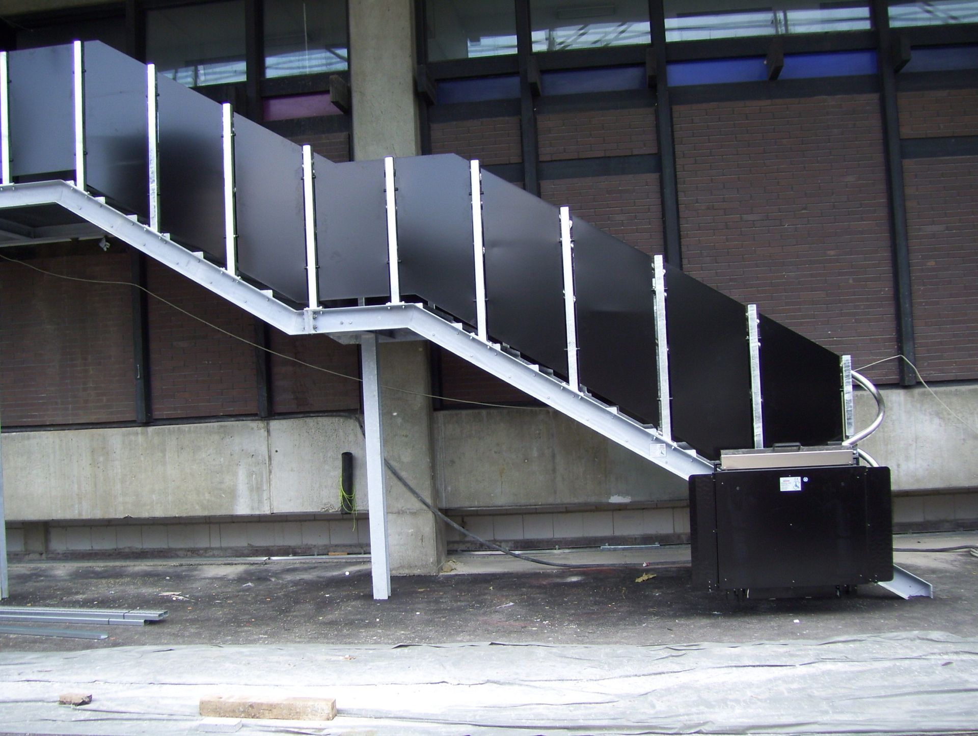 Une plateforme élévatrice noire pour fauteuil roulant est installée sur un escalier dans un couloir carrelé et lumineux, agrémenté d'un lustre.