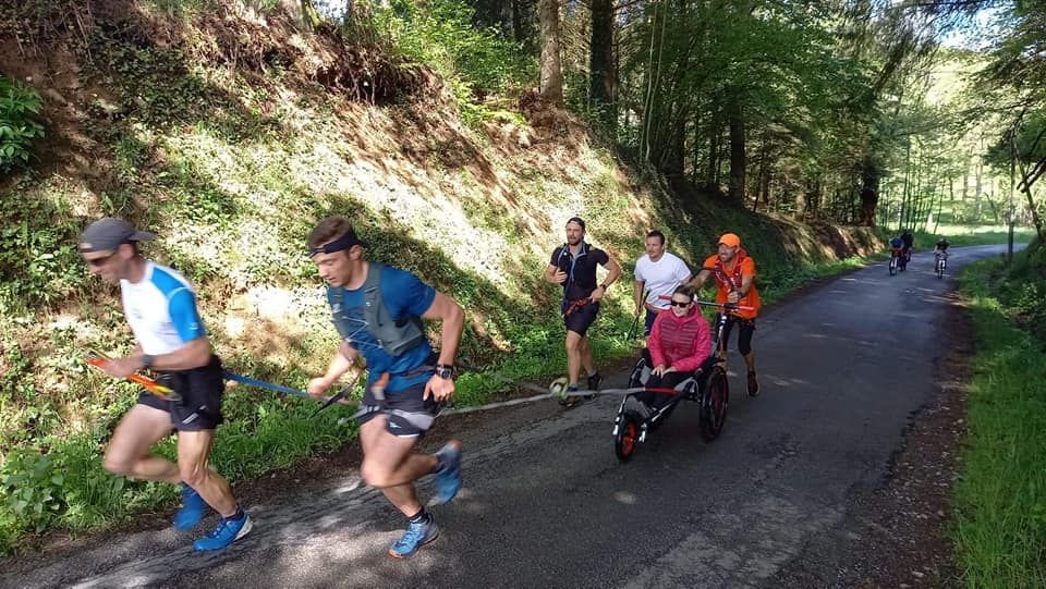 Des coureurs tirent un fauteuil roulant avec une personne à bord sur une route goudronnée traversant une zone boisée.
