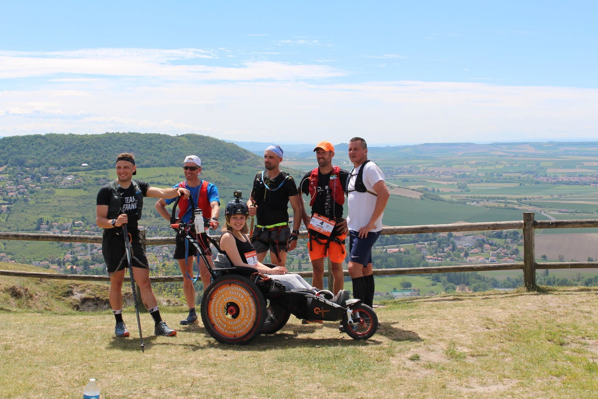 Un groupe de six personnes, dont une en fauteuil roulant, pose en extérieur devant un paysage pittoresque.