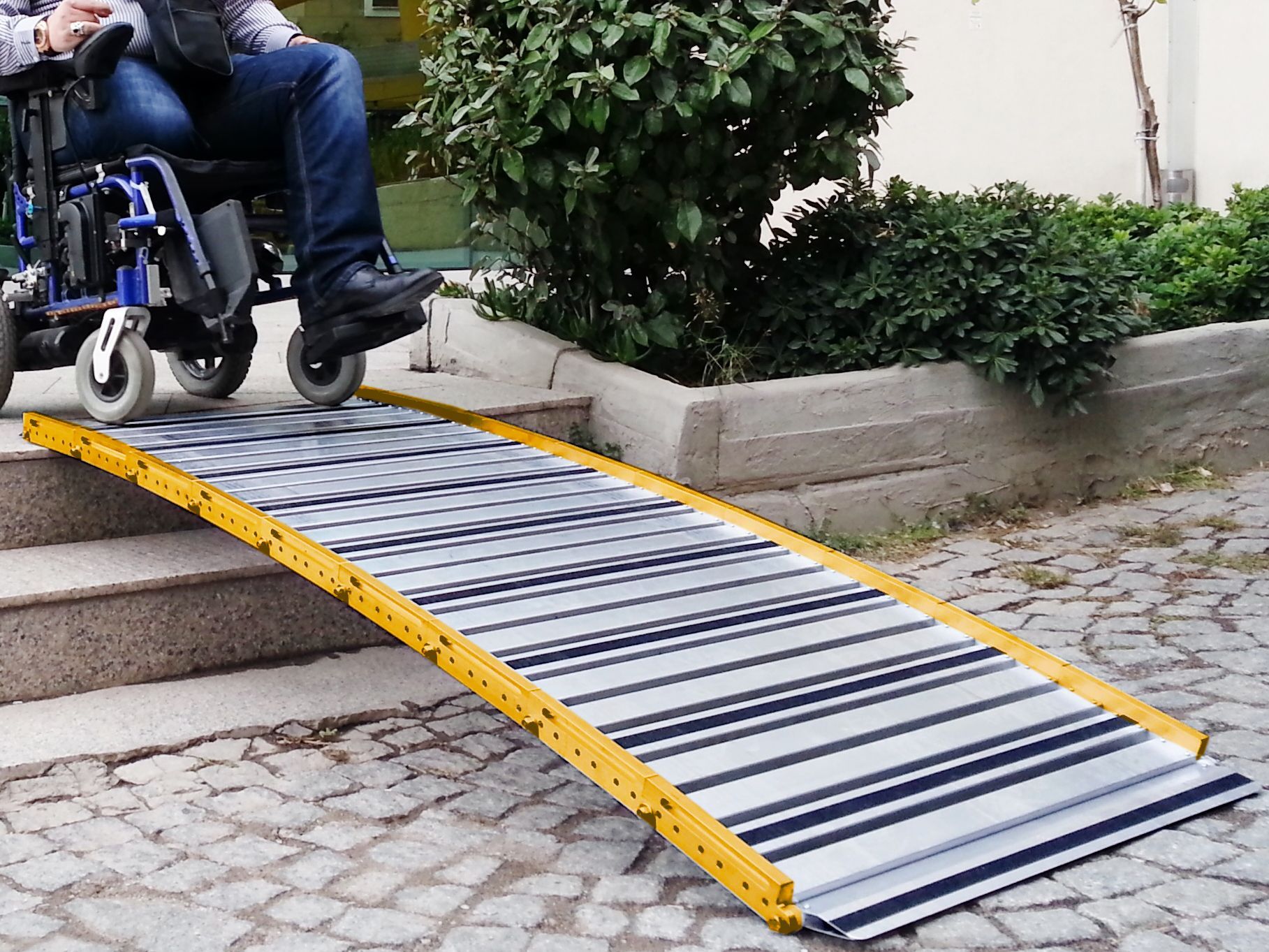 Une personne en fauteuil roulant utilise une rampe métallique portable à bord jaune pour monter des marches en pierre à l'extérieur.