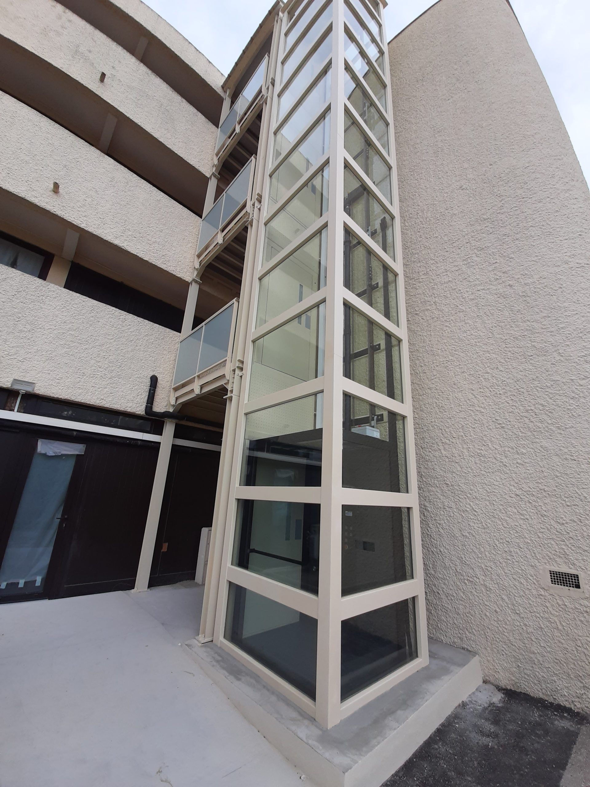 Ascenseur extérieur vitré contre un immeuble en stuc beige.