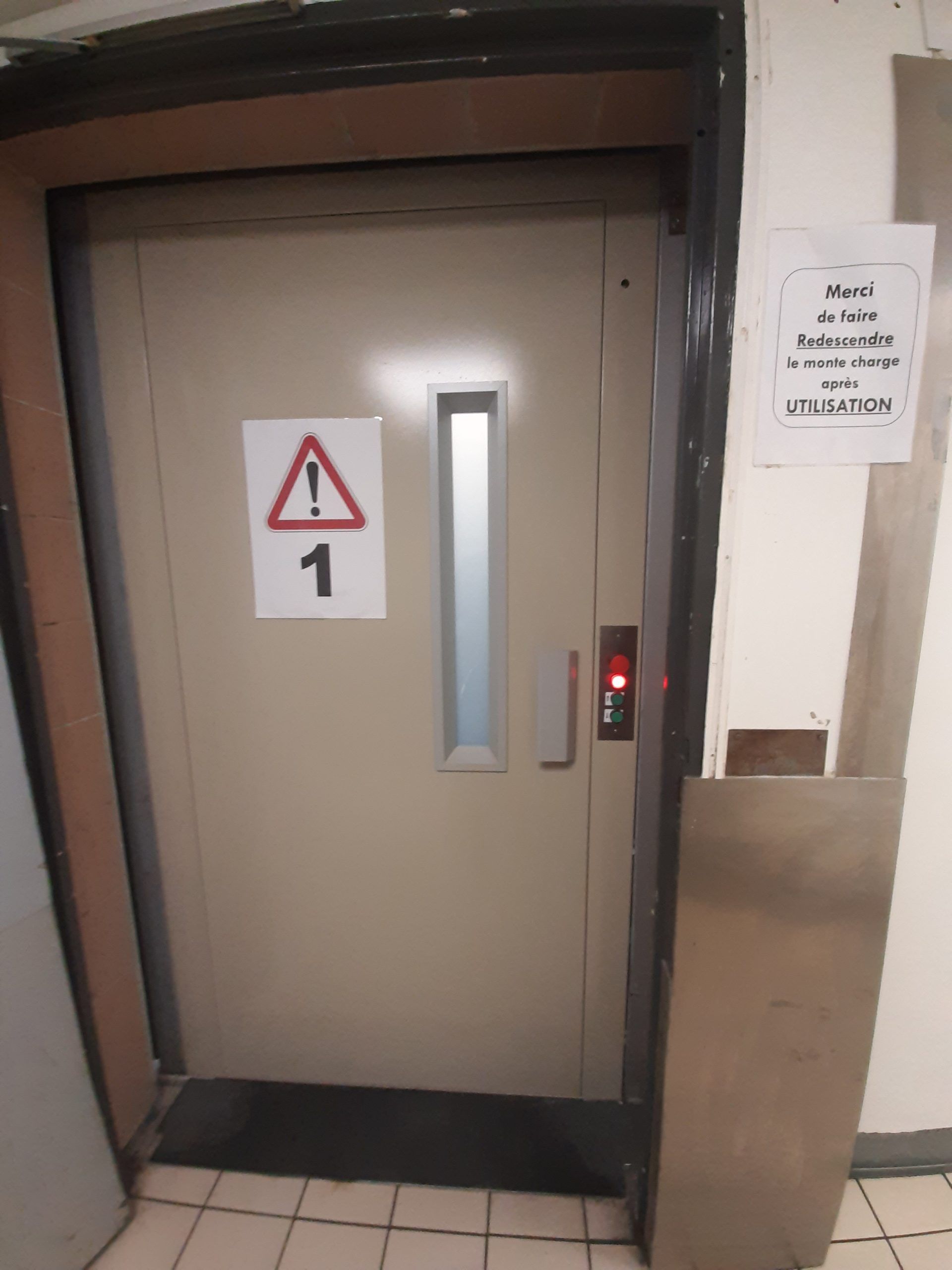Palier d'ascenseur avec porte beige fermée, fenêtre étroite et panneau d'avertissement sur un panneau métallique.