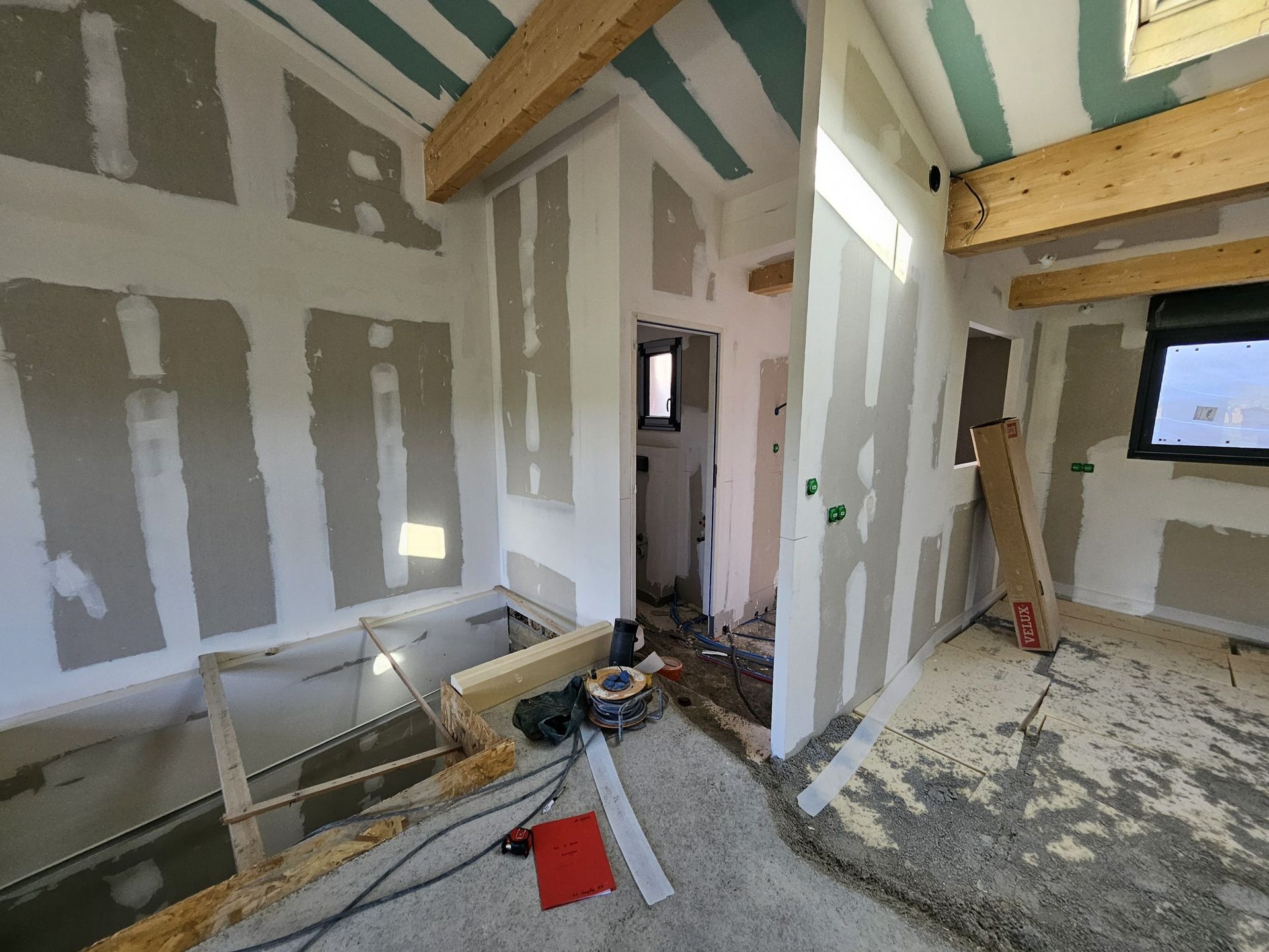 Pièce inachevée avec joints de cloisons sèches, poutres apparentes, une fenêtre et des outils de construction au sol.