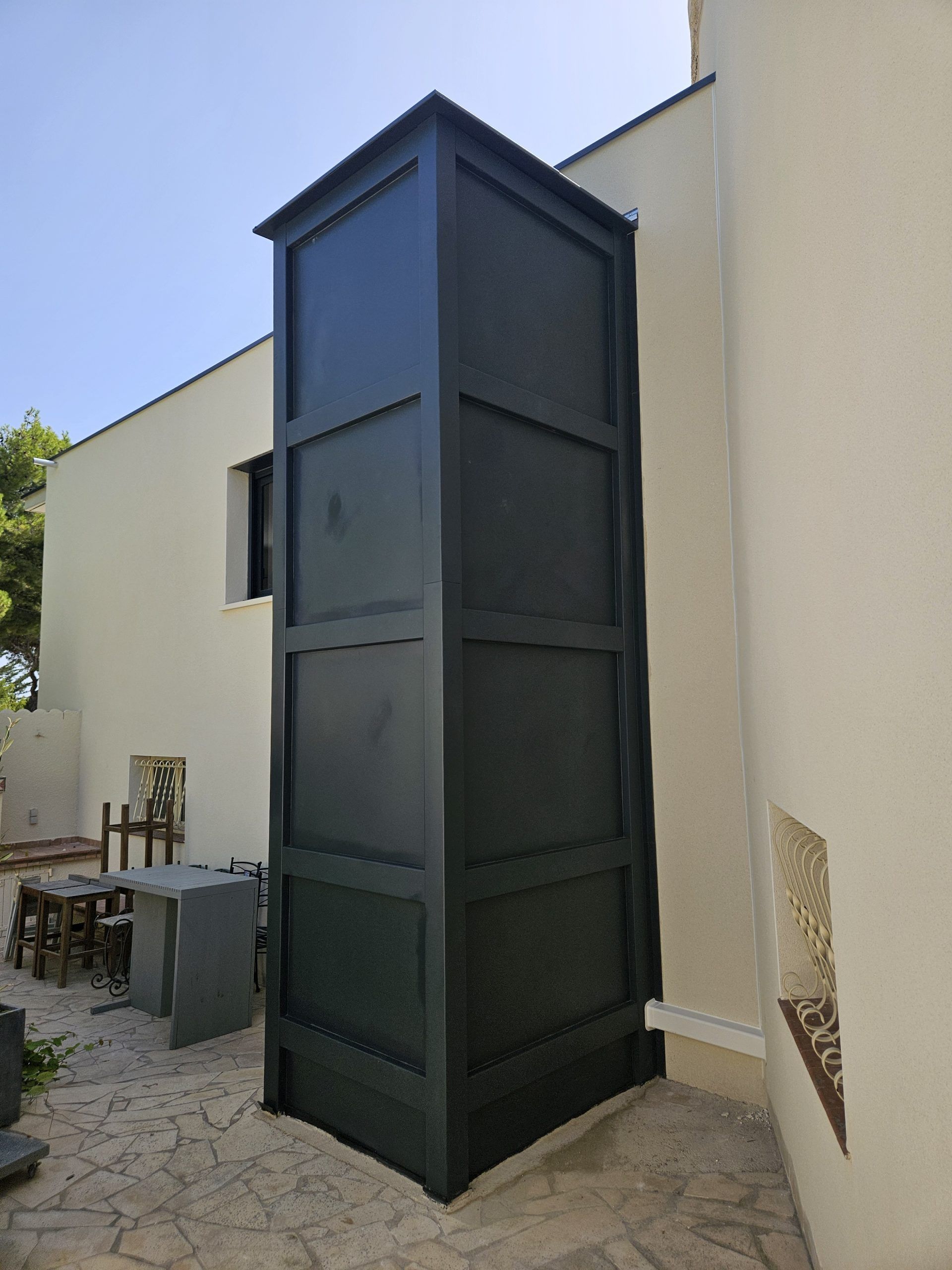Cage d'ascenseur extérieure rectangulaire noire, fixée à un bâtiment, sur une terrasse en pierre.
