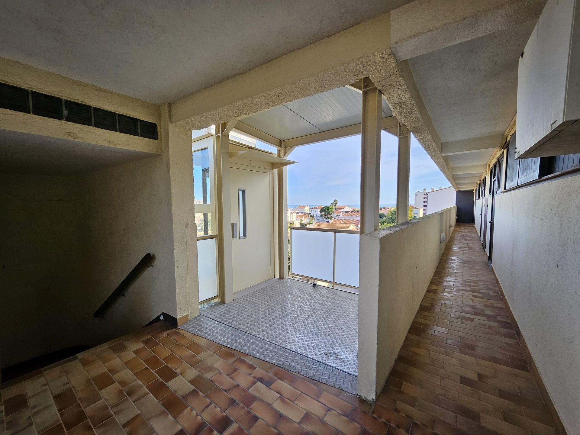Un long couloir extérieur avec un balcon ouvert donnant sur la ville. Sol en briques et poutres de soutien en béton.
