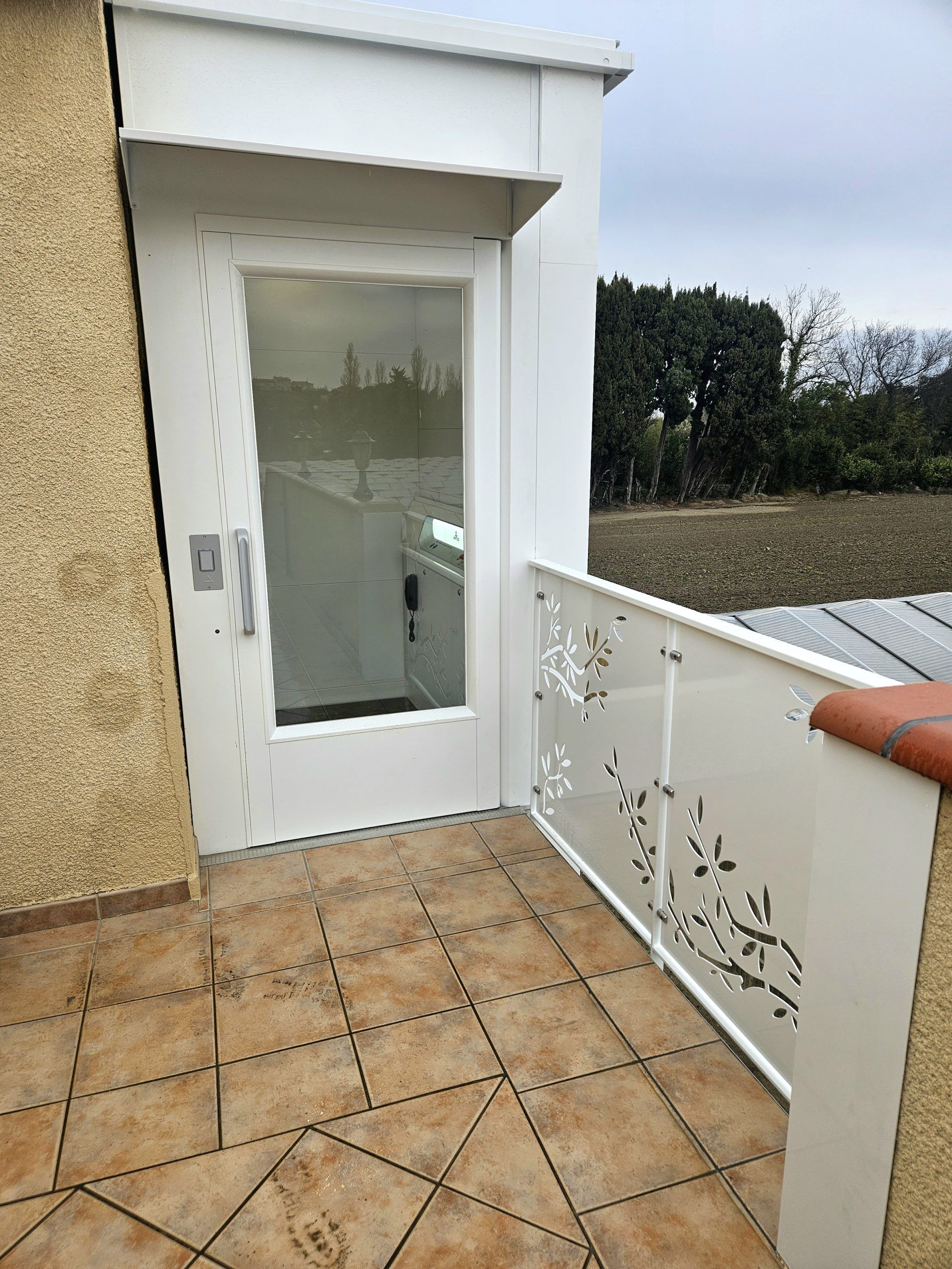 Porte blanche au bout d'une terrasse carrelée, avec une balustrade décorative et une haie à côté.
