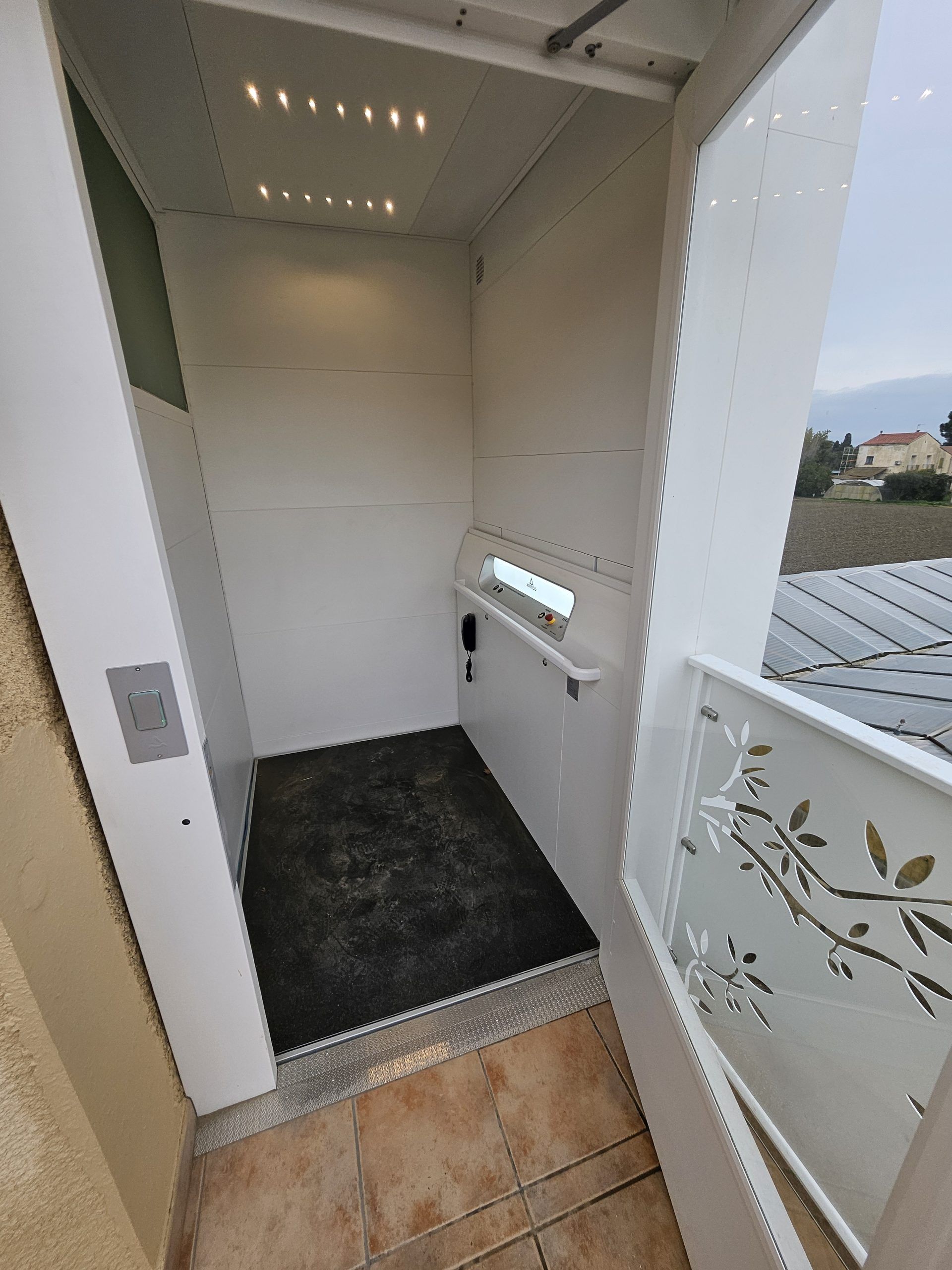 Intérieur d'ascenseur blanc de petite taille, avec tapis de sol noir et porte en verre dépoli ouverte.
