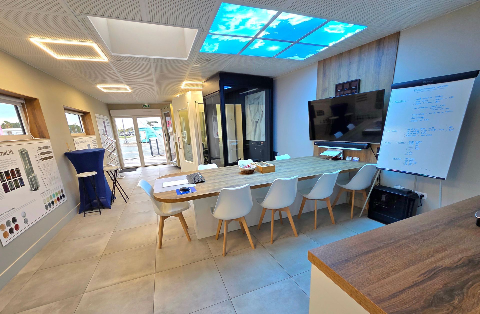 Une salle de réunion moderne comprend une table centrale en bois, des chaises blanches, un grand écran mural et un chevalet blanc.