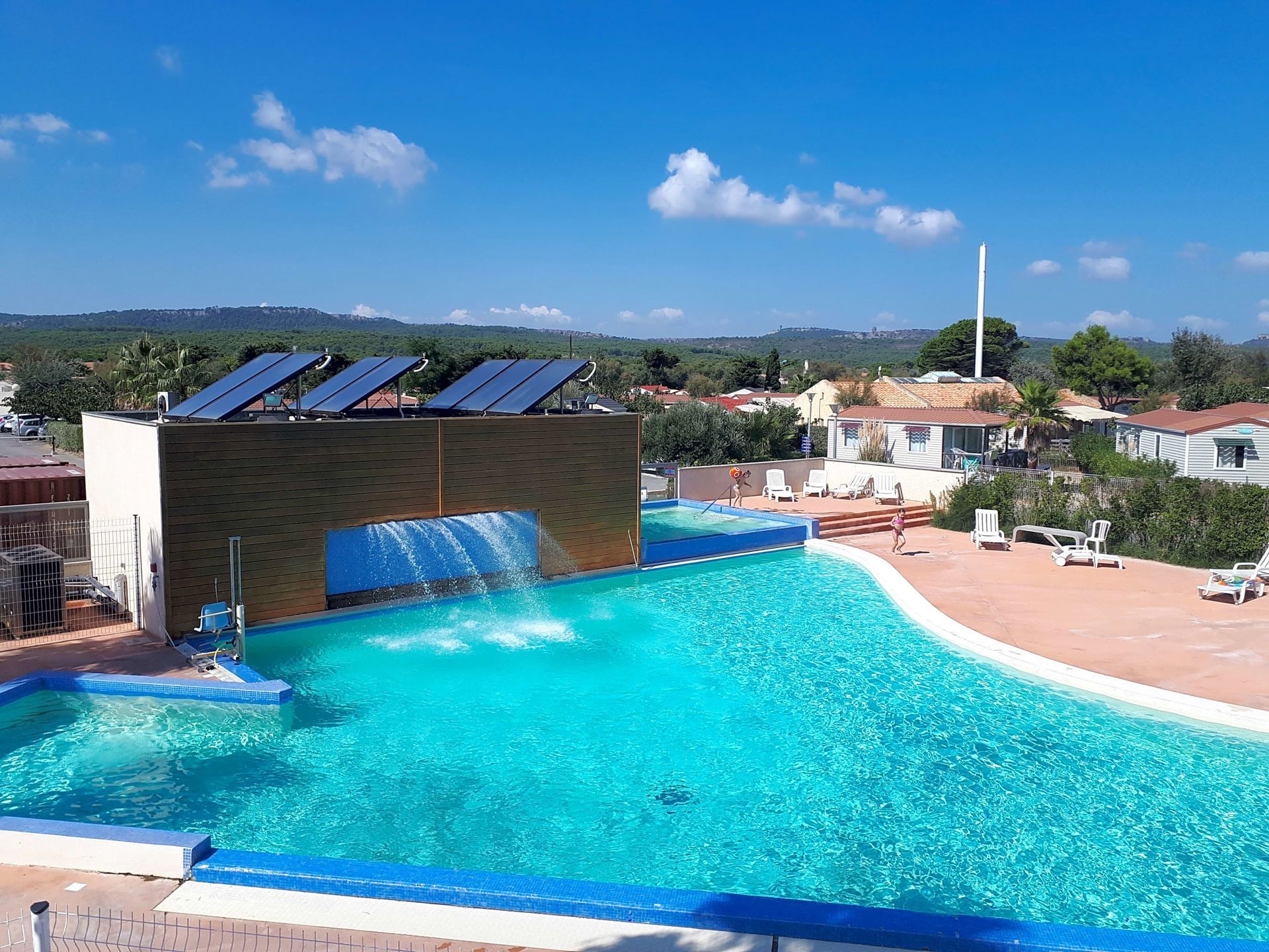 Piscine avec cascade, entourée de chaises longues sous un ciel bleu. Panneaux solaires sur un mur.