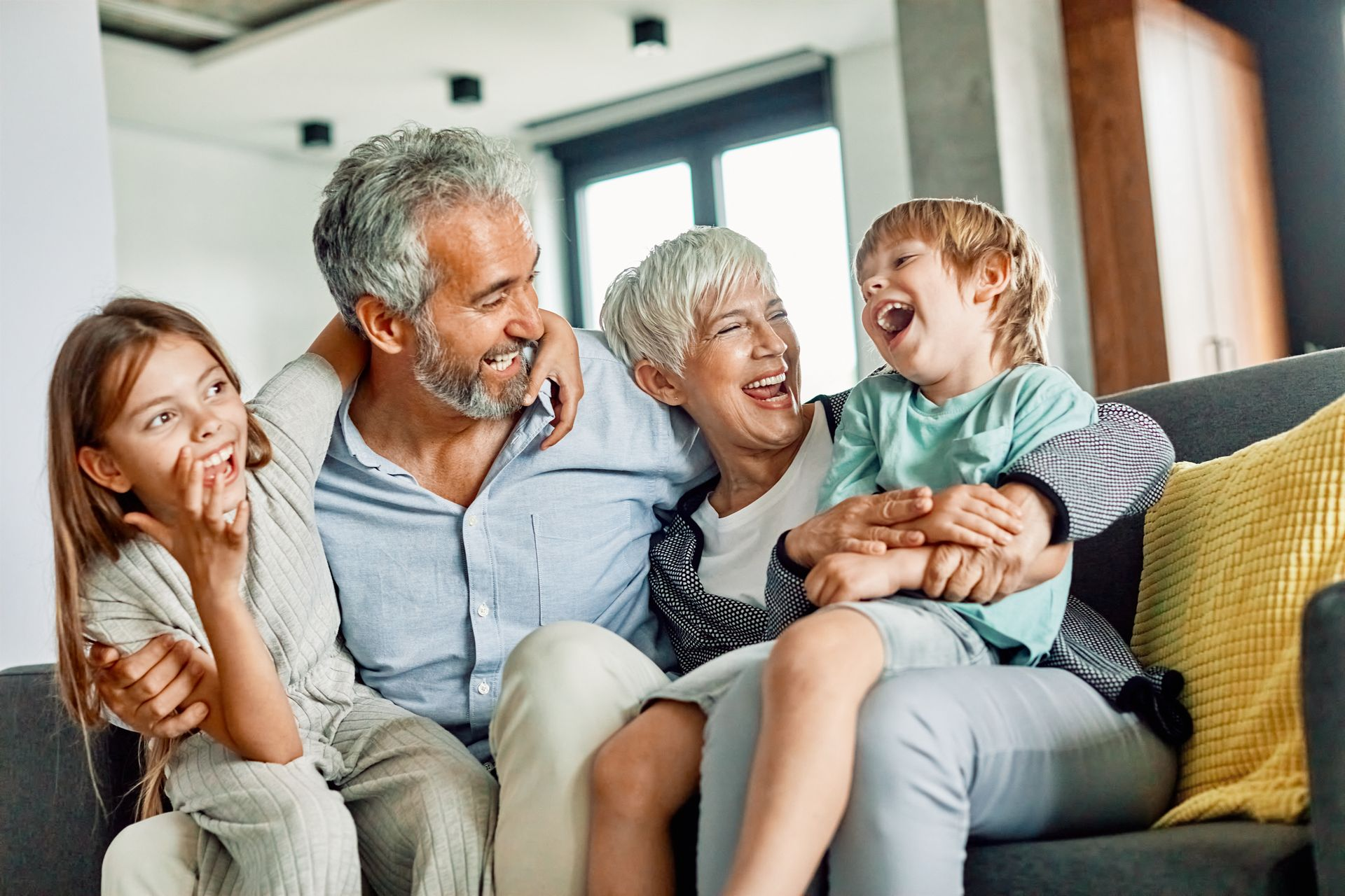 Grands-parents et petits-enfants rient ensemble sur un canapé ; fenêtre ensoleillée en arrière-plan.