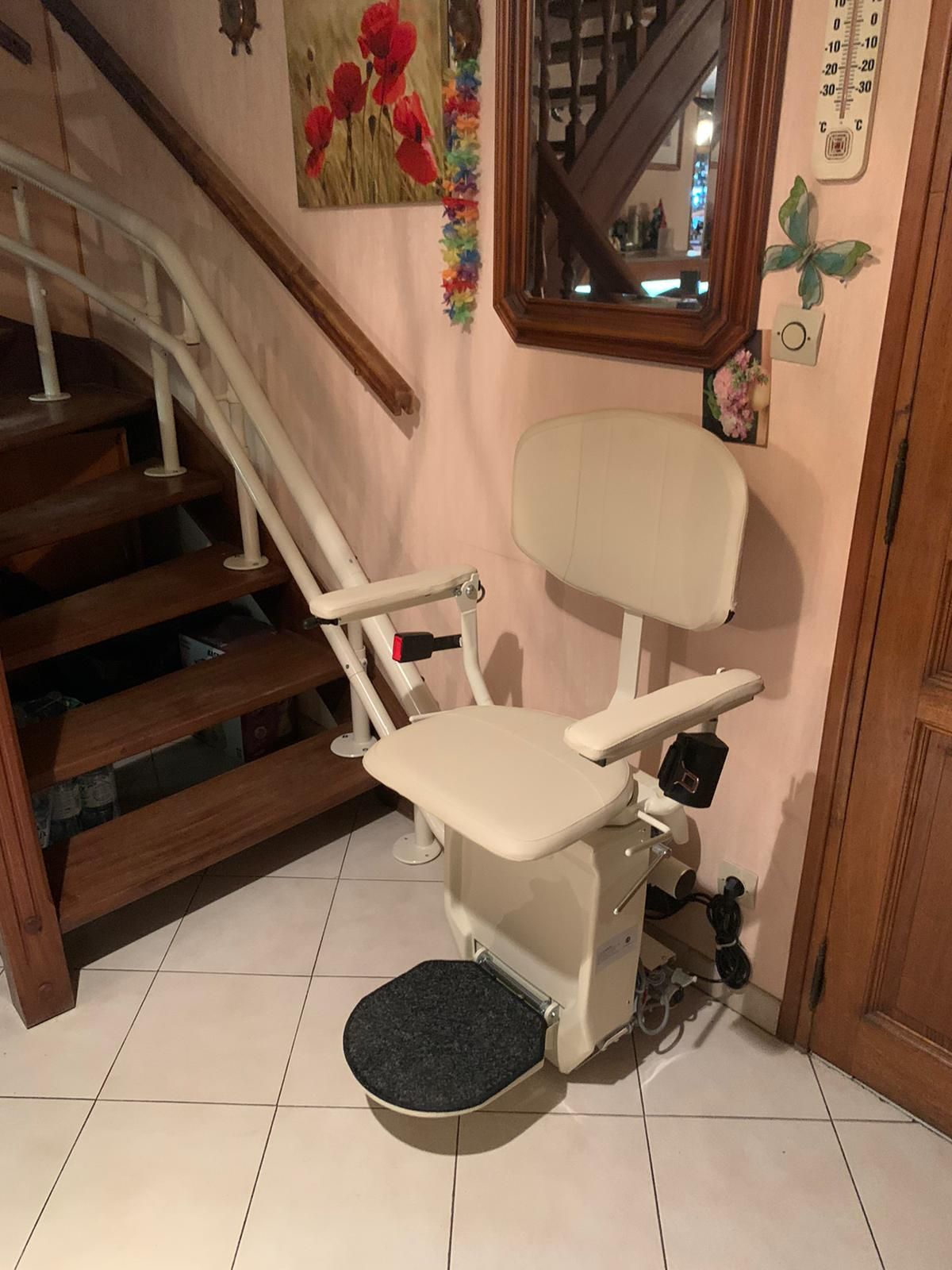 Fauteuil monte-escalier sur un escalier. Fauteuil beige avec accoudoirs et repose-pieds gris. Marches marron et rampe en bois.