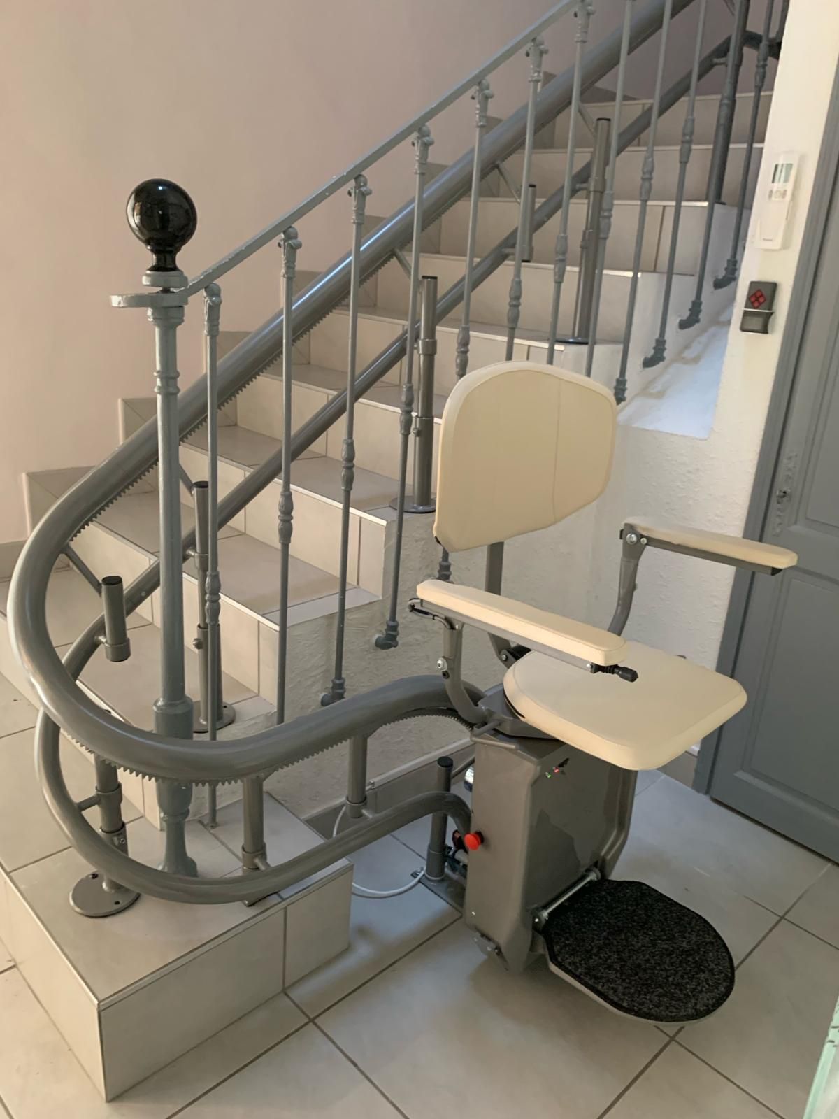 Un monte-escalier est installé sur un escalier courbe avec une rampe grise et des marches blanches. Le siège du monte-escalier est de couleur crème.