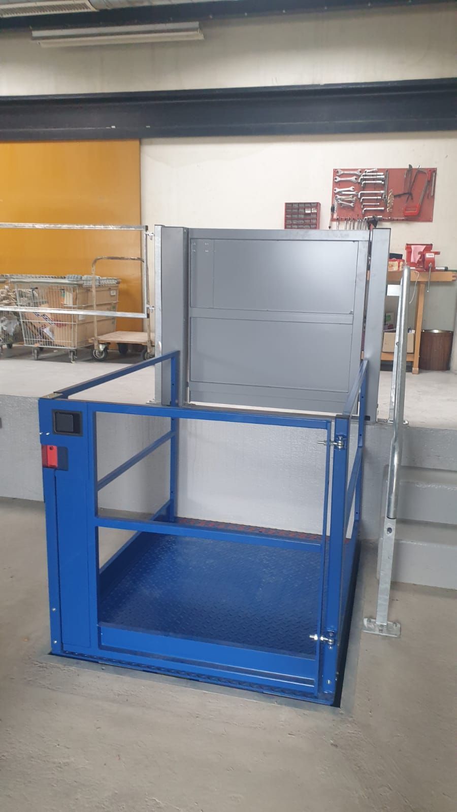 Cage de sécurité métallique bleue dans un atelier, à côté d'une machine industrielle grise et d'un escalier.