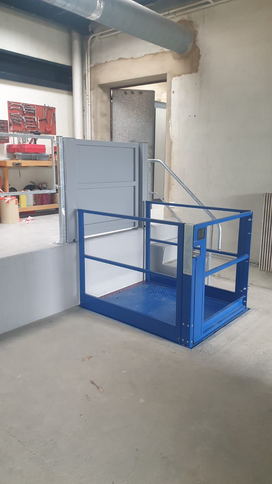 Plateforme élévatrice bleue et grise avec garde-corps, située à côté d'une porte, dans un entrepôt.
