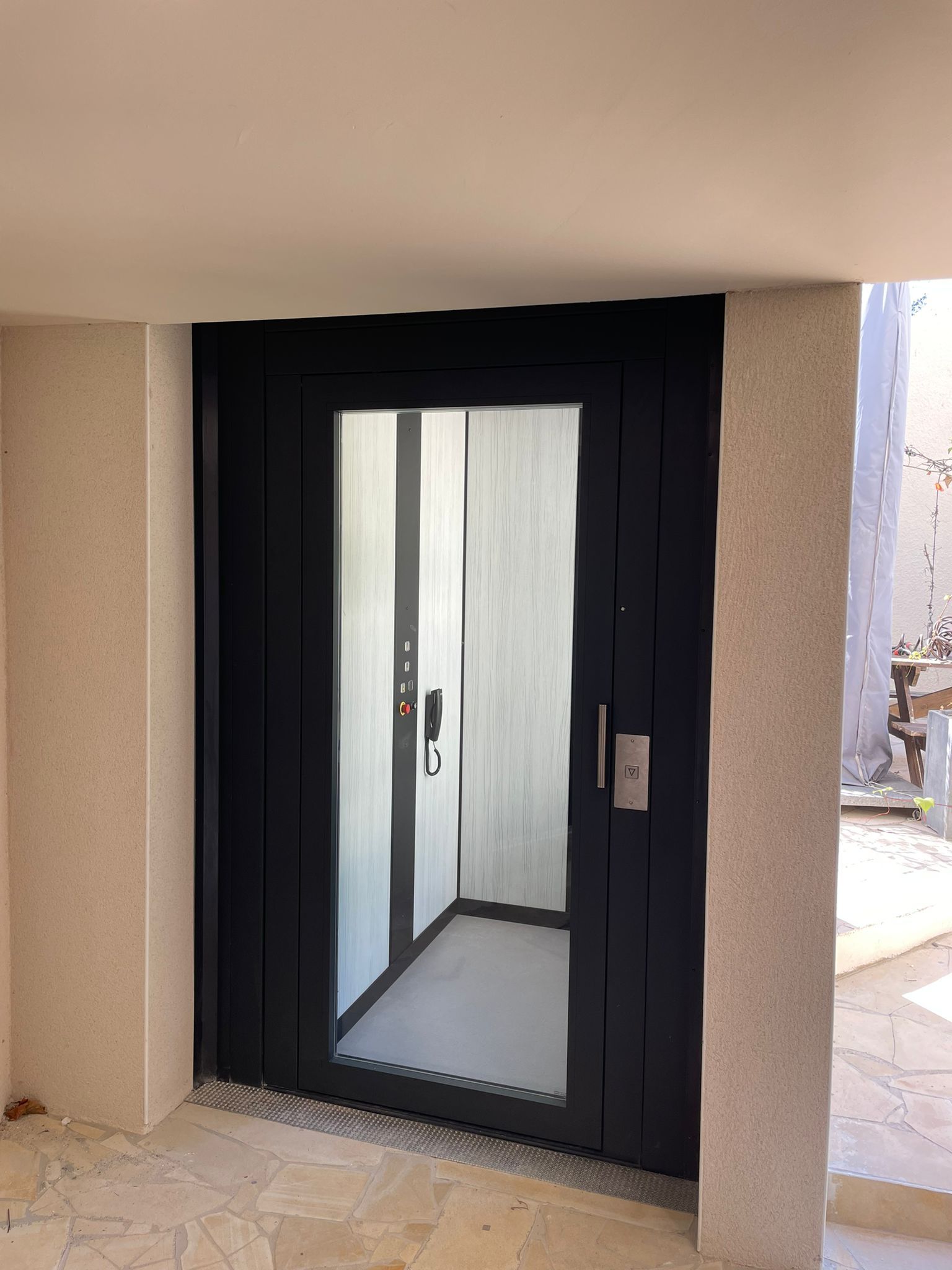 Porte d'ascenseur noire avec panneau de verre et poignée, entre des murs beiges.