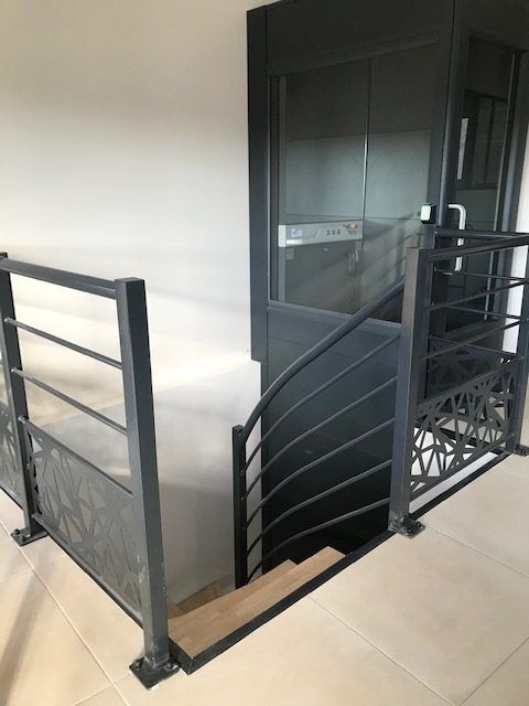 Escalier avec rampe en métal noir et accès par ascenseur ; espace d'entrée avec murs et sol clairs.