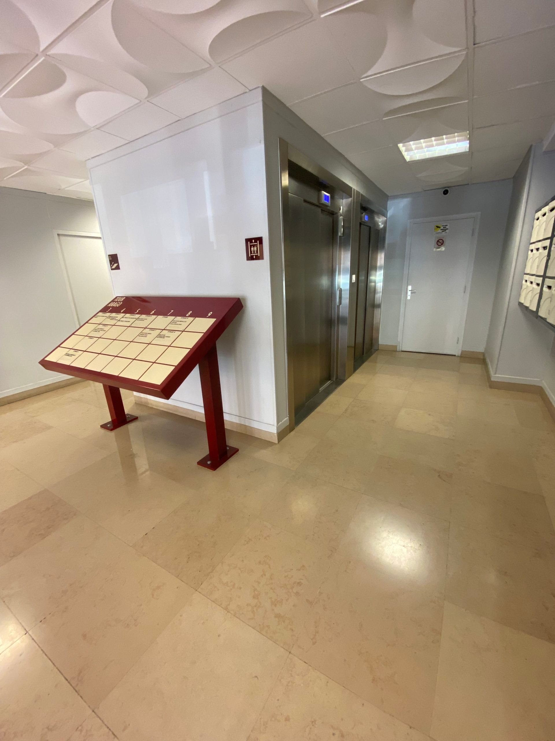 Couloir de bureau avec portes d'ascenseur, murs blancs et présentoir rouge sur un sol beige.