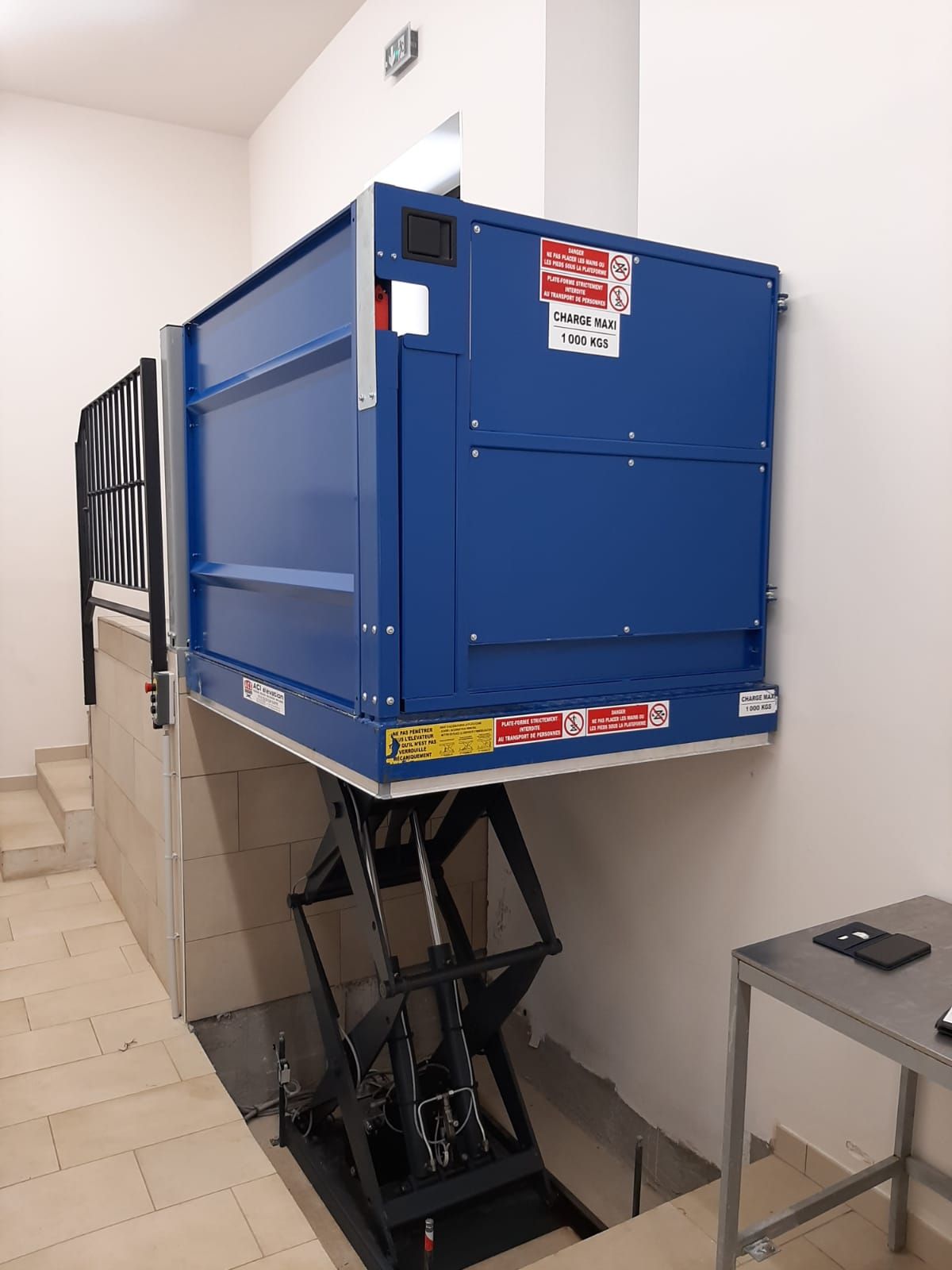 Un monte-charge bleu est installé au-dessus d'un escalier. Il repose sur un mécanisme à ciseaux noir.