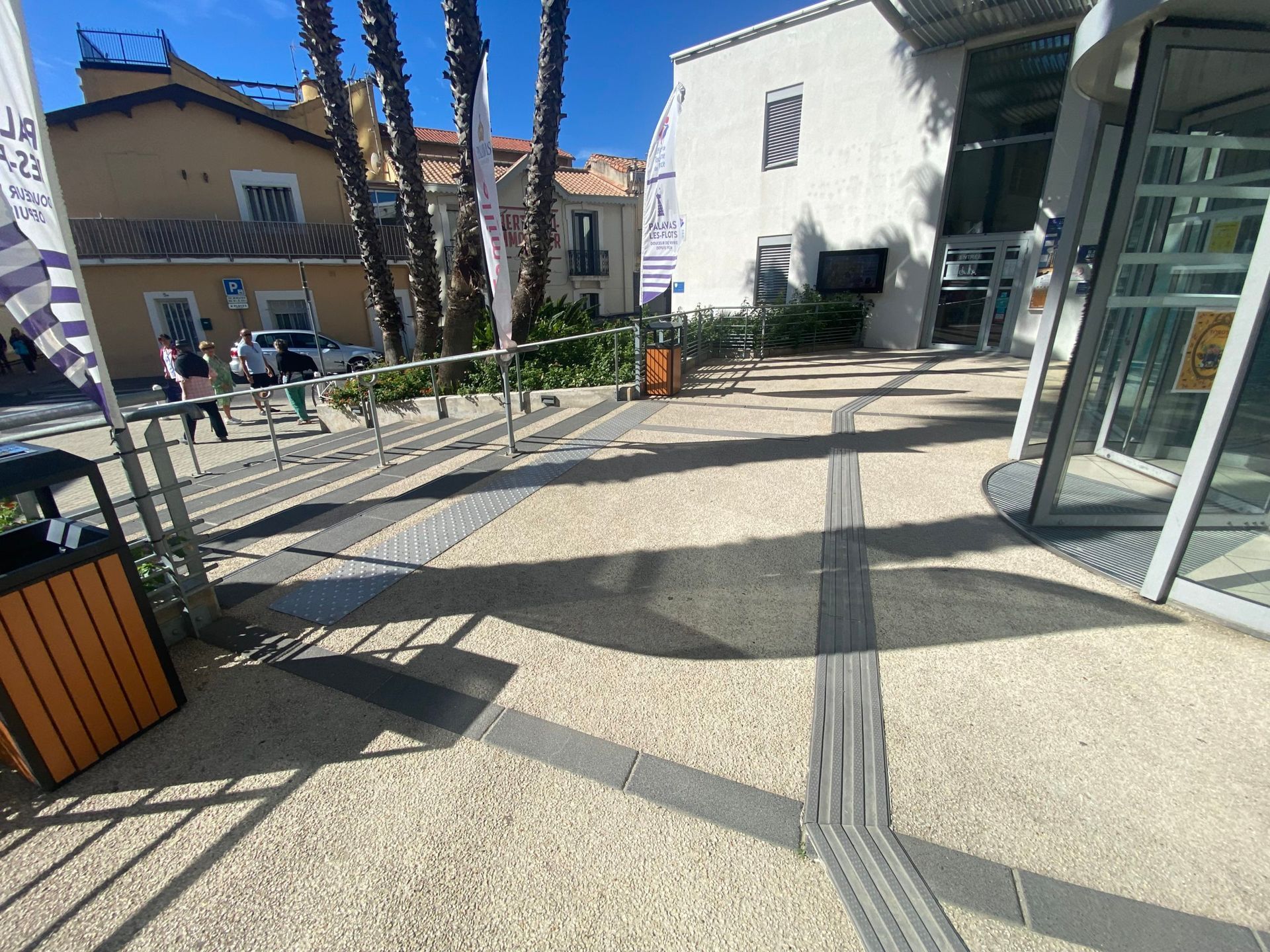 Extérieur d'un bâtiment avec des voies d'accès accessibles balisées par des bandes texturées. Présence d'une rampe et d'escaliers.