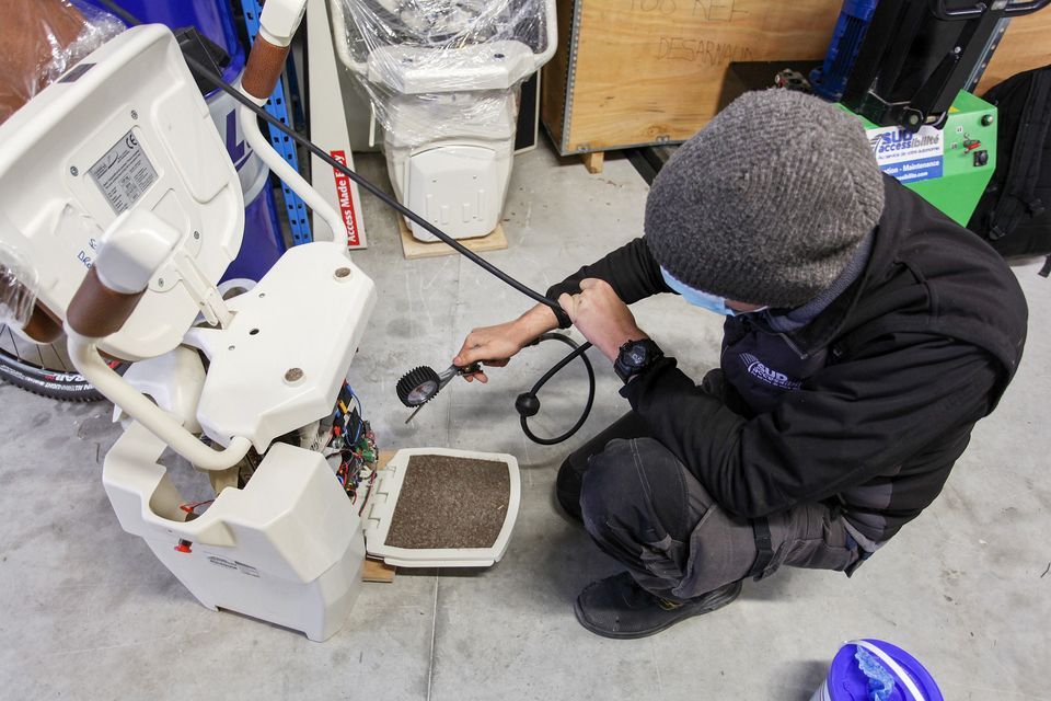 Un technicien portant un bonnet gris s'accroupit pour inspecter la base ouverte d'un monte-escalier blanc dans un entrepôt.