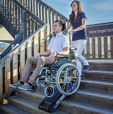 Un homme en fauteuil roulant monte un escalier, aidé par une femme munie d'un monte-escalier portatif. Escalier en bois, journée ensoleillée.