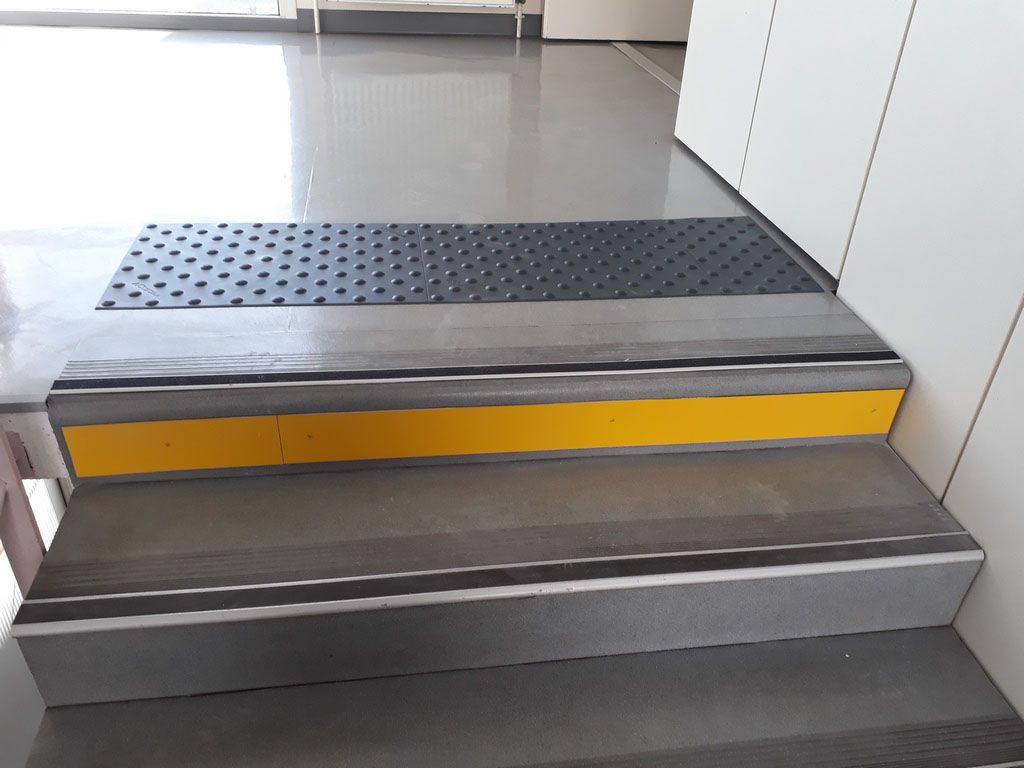 Quelques marches métalliques courtes avec un bord jaune et un tapis tactile noir à l'entrée.