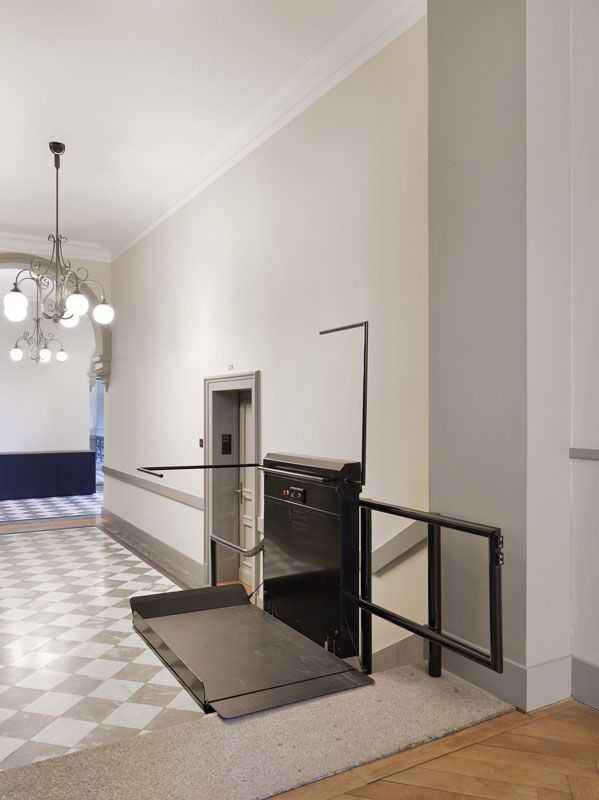 Une plateforme élévatrice noire pour fauteuil roulant est installée sur un escalier dans un couloir carrelé et lumineux, agrémenté d'un lustre.
