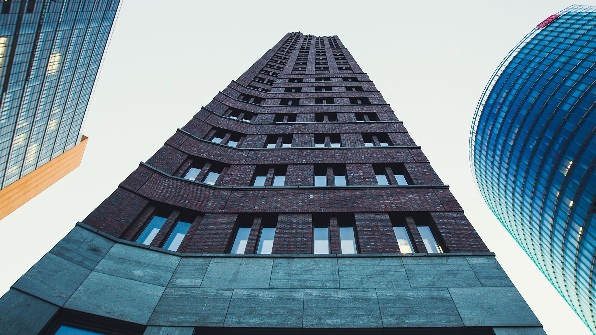 Vista desde un ángulo bajo del edificio Flatiron de ladrillo oscuro en Berlín, flanqueado por modernos rascacielos de cristal contra un cielo brillante.