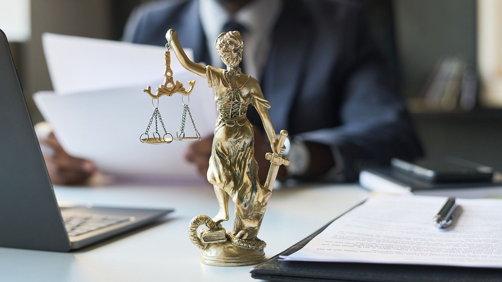 Una estatua dorada de la Dama de la Justicia reposa sobre un escritorio frente a un profesional que trabaja con papeles y una computadora portátil.