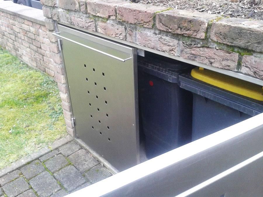 Unter einer Backsteinmauer steht ein Mülleimer aus Edelstahl.
