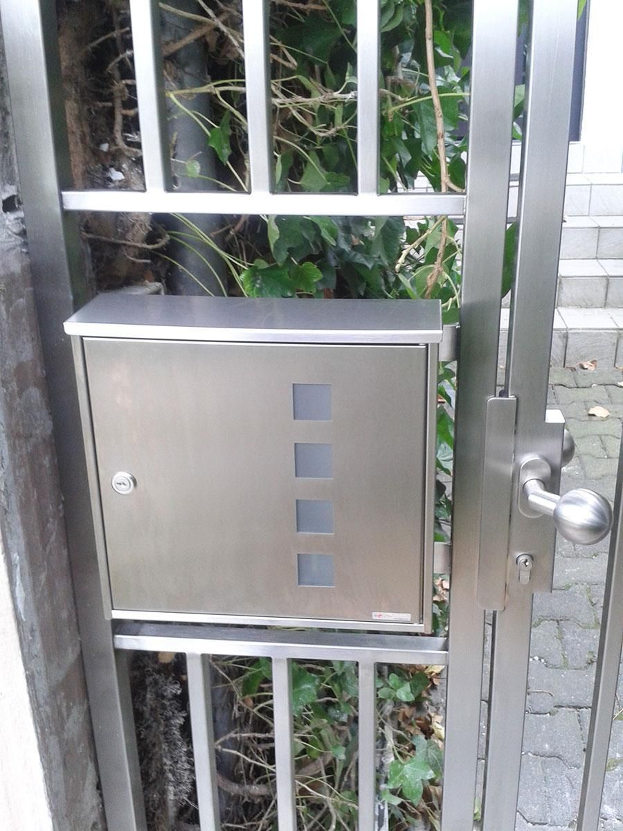 An einem Metalltor ist ein Briefkasten aus Edelstahl angebracht.