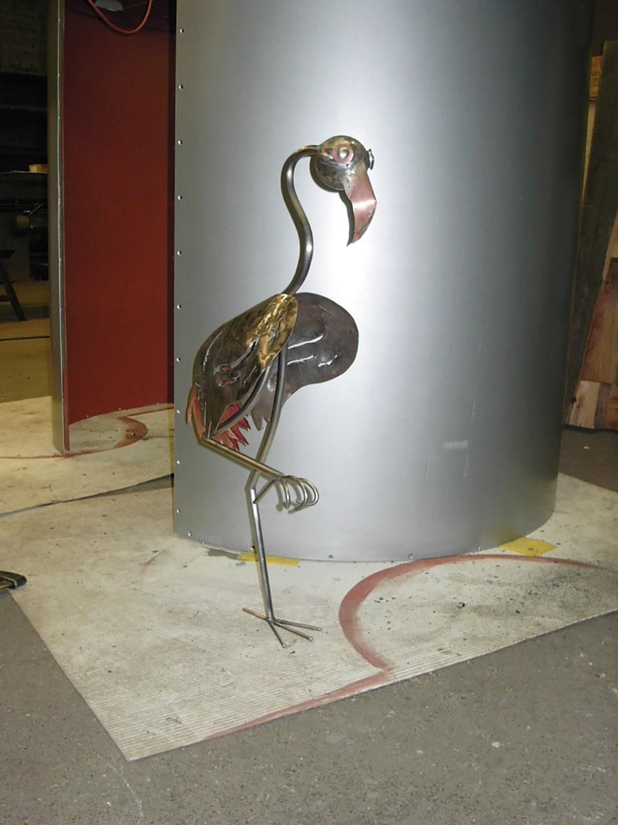 Eine Flamingostatue aus Metall steht neben einem Metallzylinder.