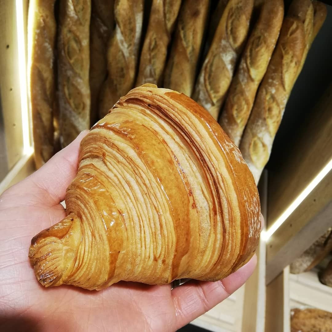 Une main tenant un croissant doré et feuilleté devant une étagère de baguettes.