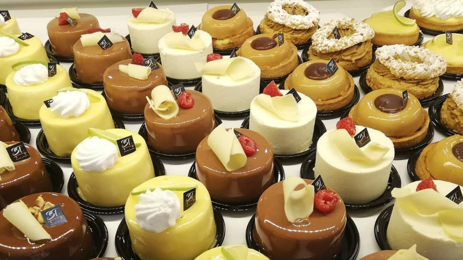 Des rangées de gâteaux miniatures aux glaçages colorés et aux saveurs assorties, présentés dans une vitrine de boulangerie.
