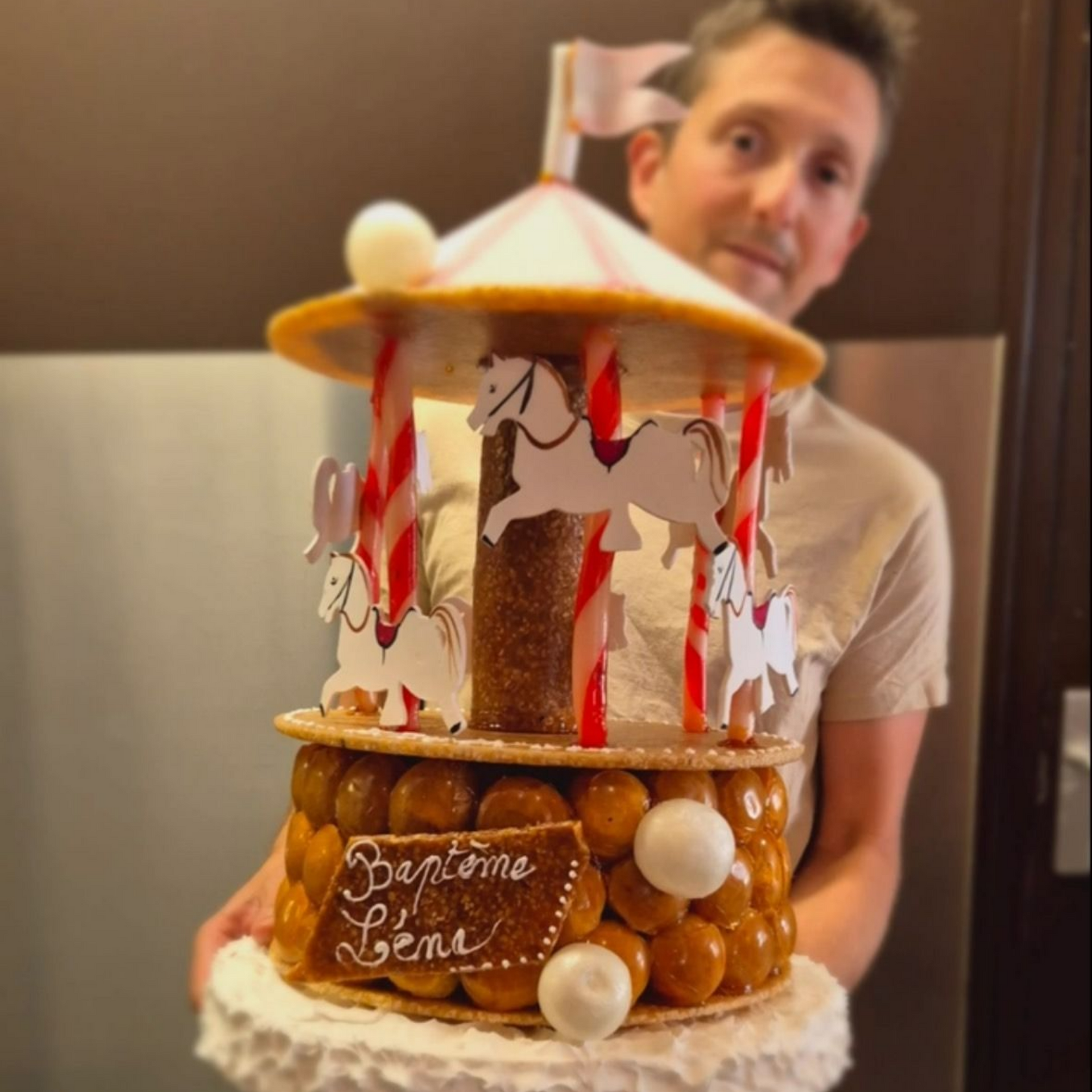 Un homme tient un gâteau en forme de carrousel. Le gâteau est brun, blanc et rouge, avec un symbole de baptême.