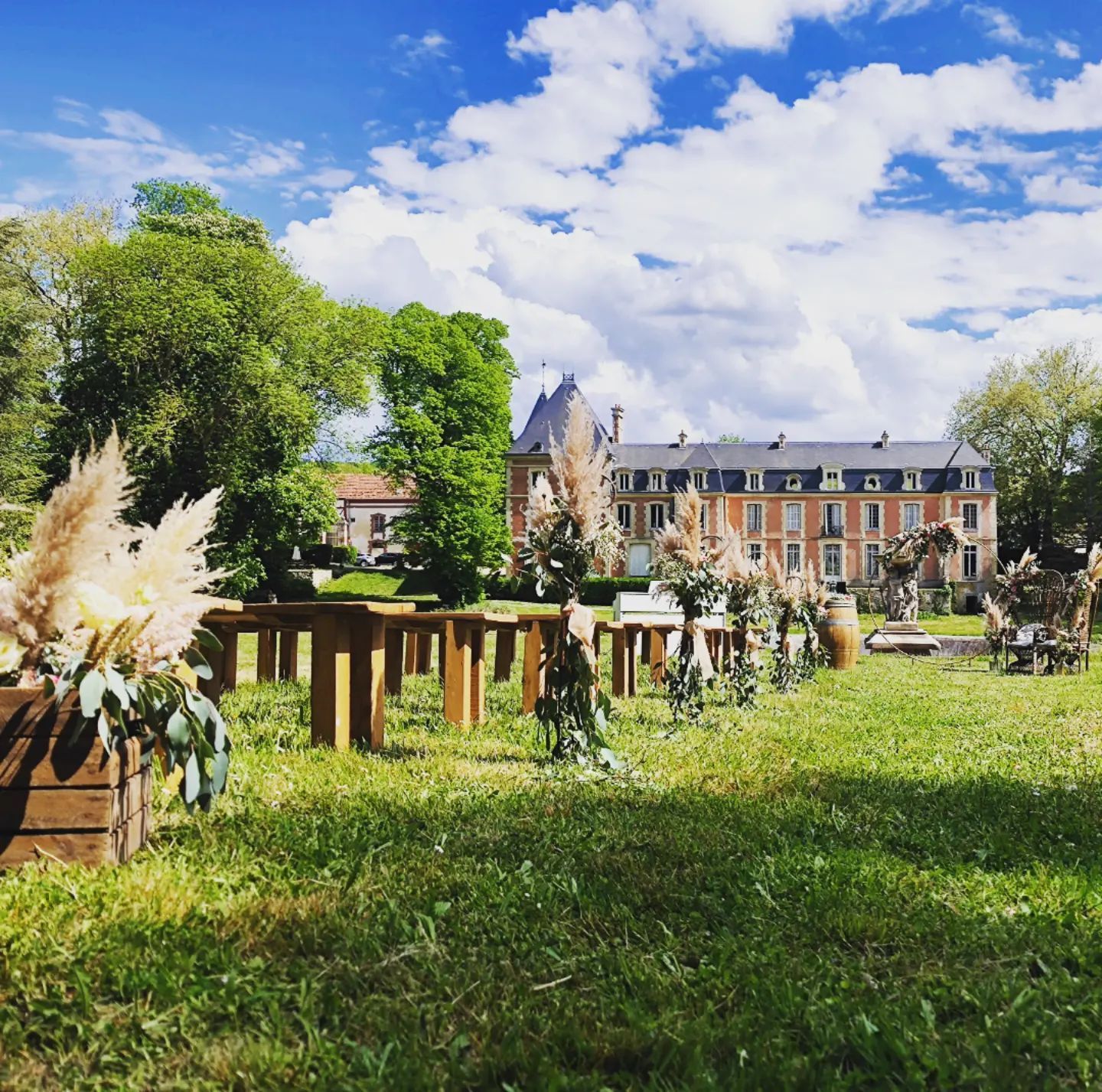 Jardin ensoleillé avec des fleurs, des bancs en bois et un grand bâtiment historique sous un ciel bleu.