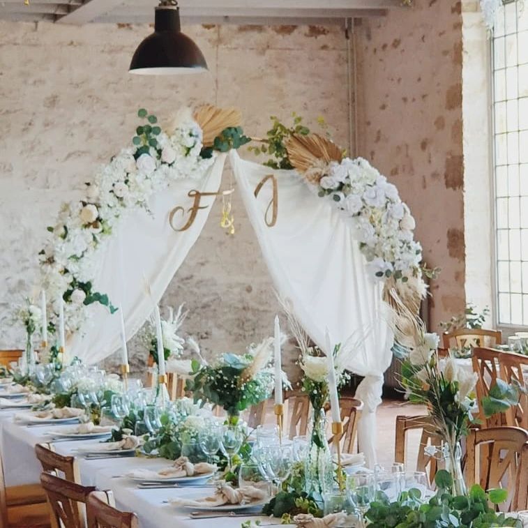 Élégante table de réception de mariage avec arche florale blanche, tissu drapé et décor rustique dans une salle lumineuse.