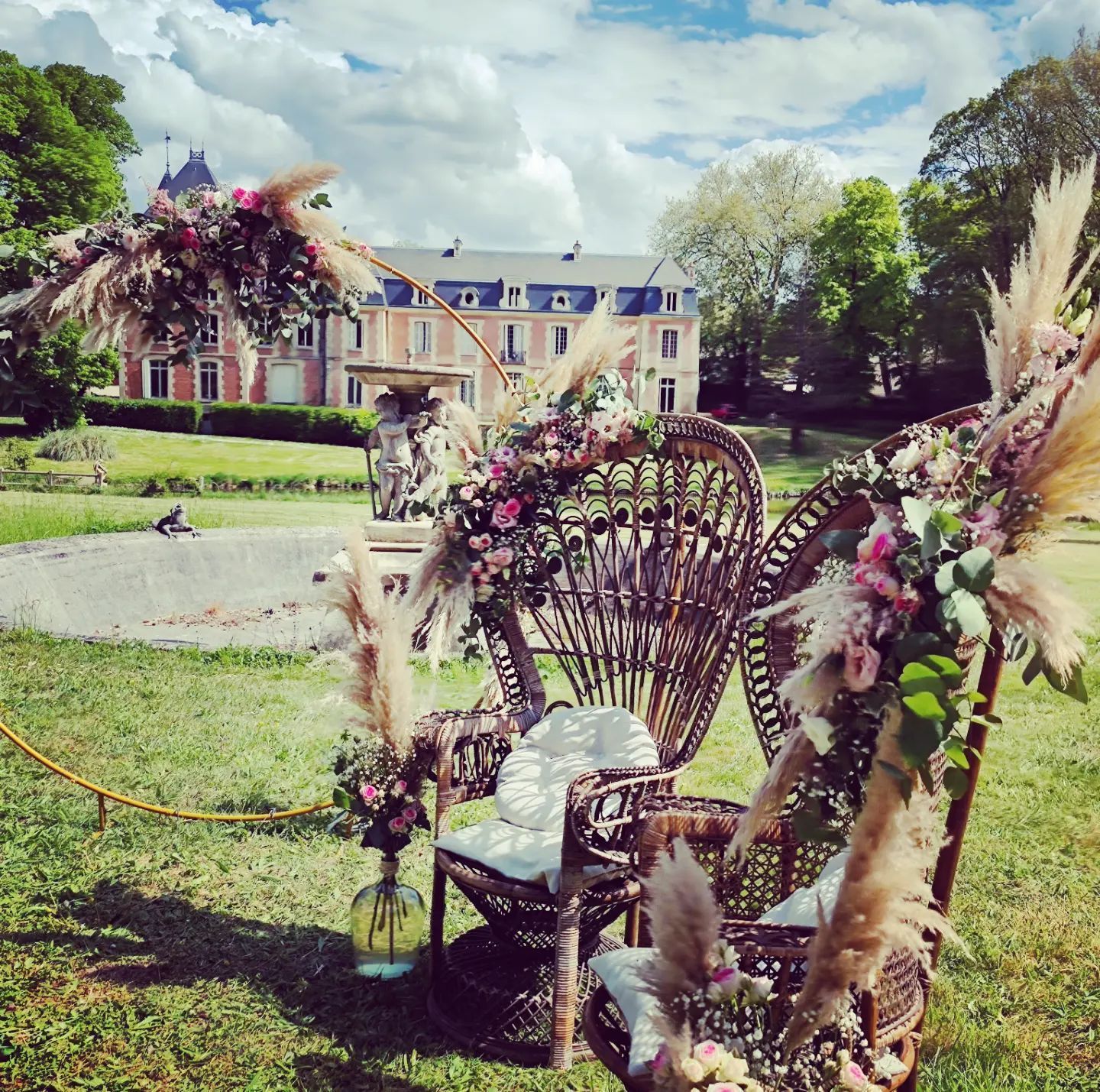 Fauteuils décoratifs en osier ornés de fleurs roses sur une pelouse devant une grande demeure sous un ciel nuageux.