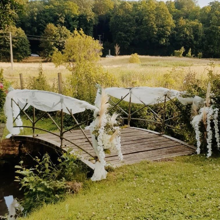 Petit pont en bois enjambant un ruisseau, drapé de tissu blanc et de fleurs dans une prairie ensoleillée.
