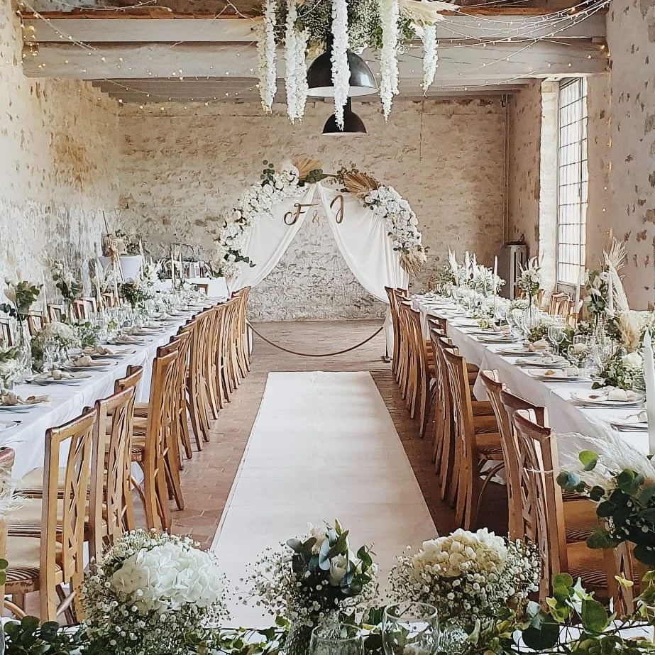 Réception de mariage champêtre et élégante avec de longues tables blanches, de la verdure et une arche fleurie.