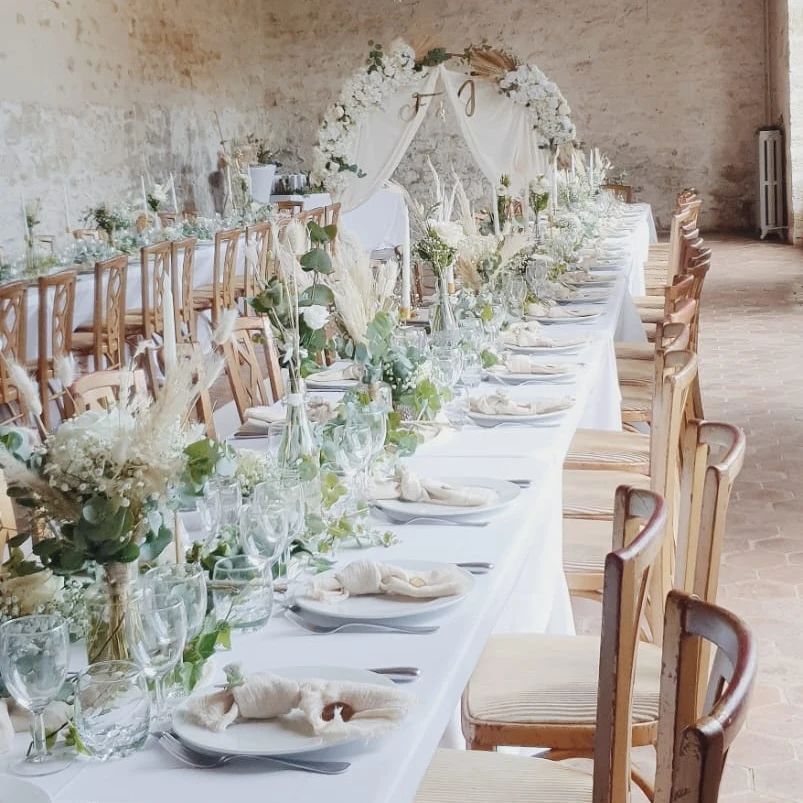 Élégante table de mariage dressée avec des nappes blanches, des fleurs et des chaises en bois dans une salle rustique.