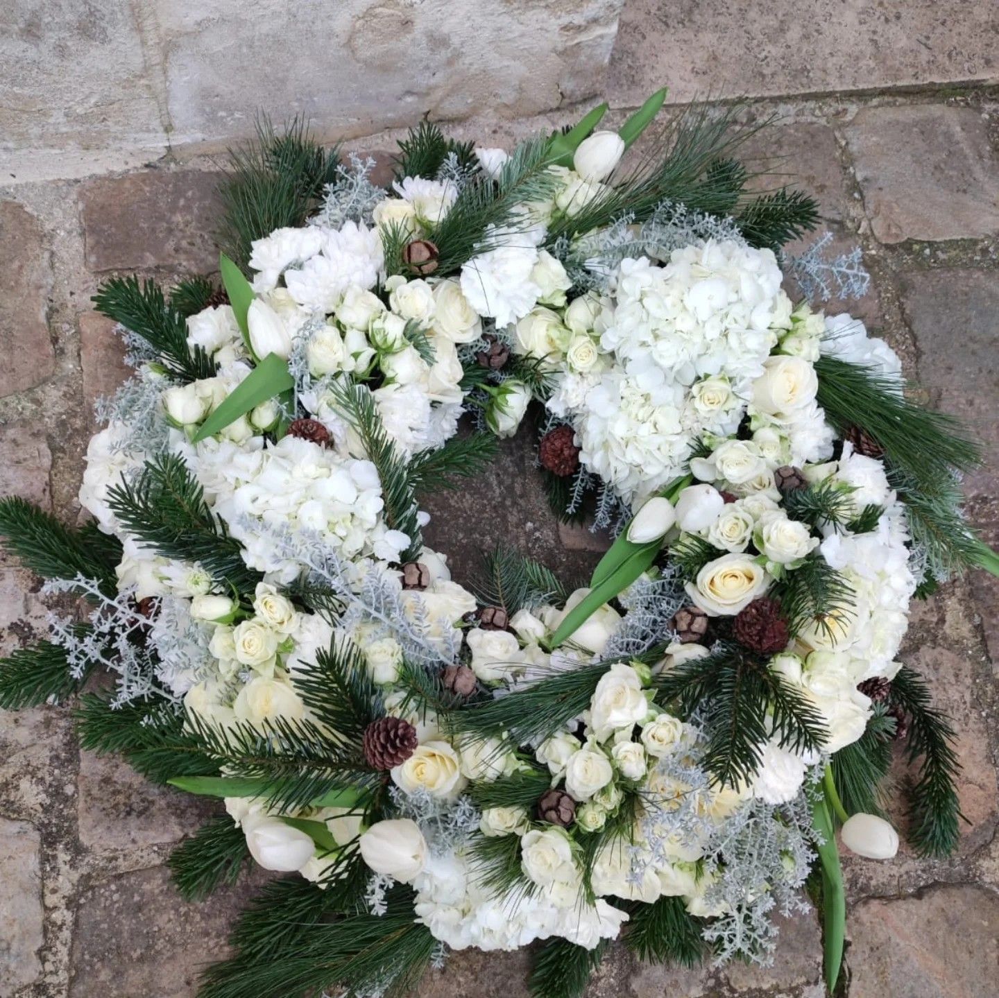 Couronne de fleurs blanches avec feuillage vert et pommes de pin sur un pavé.