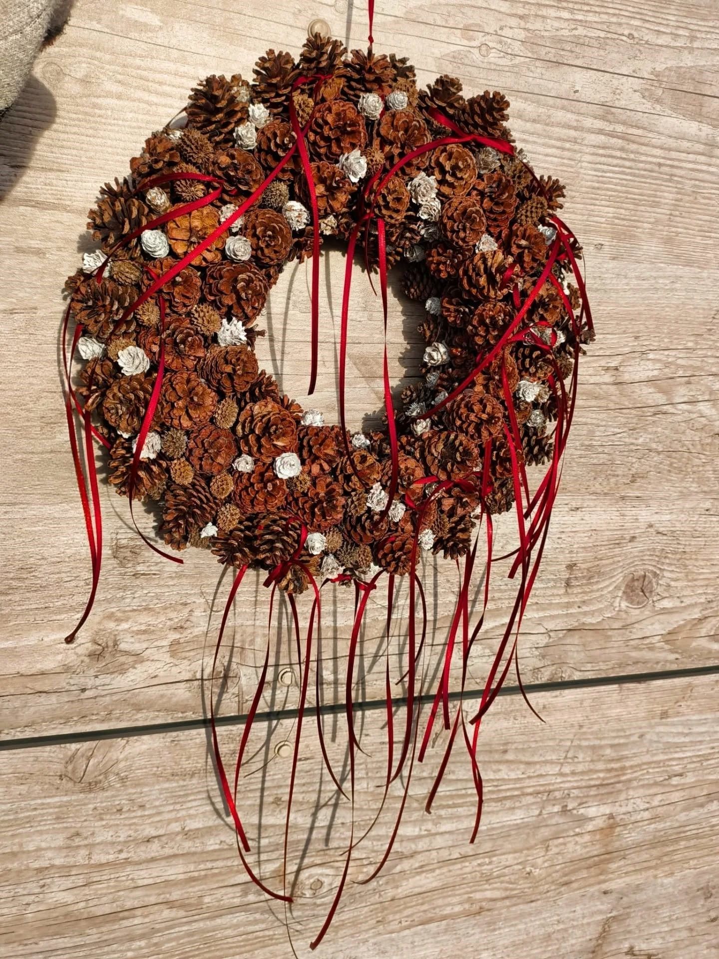 Couronne de pommes de pin brunes ornée de rubans rouges, accrochée à un mur en bois clair.
