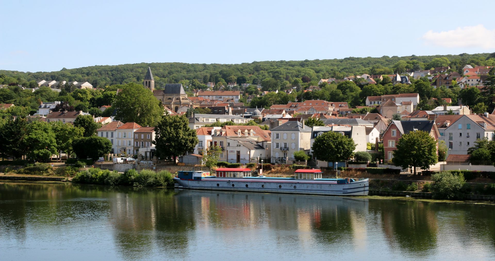Village riverain aux maisons colorées et un bateau se reflétant dans l'eau calme sous un ciel clair.