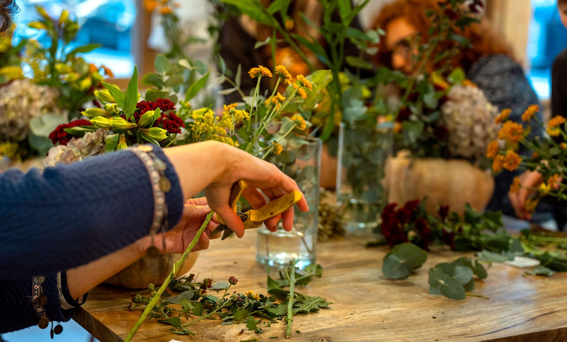 Main déposant une petite bougie en verre parmi des fleurs et de la verdure sur une table rustique.
