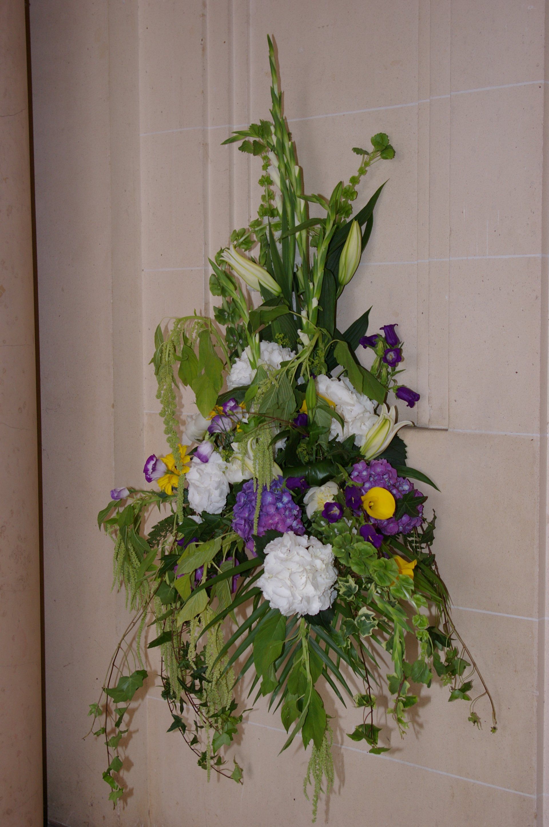 Haute composition florale avec des fleurs blanches, violettes et jaunes sur un mur beige.
