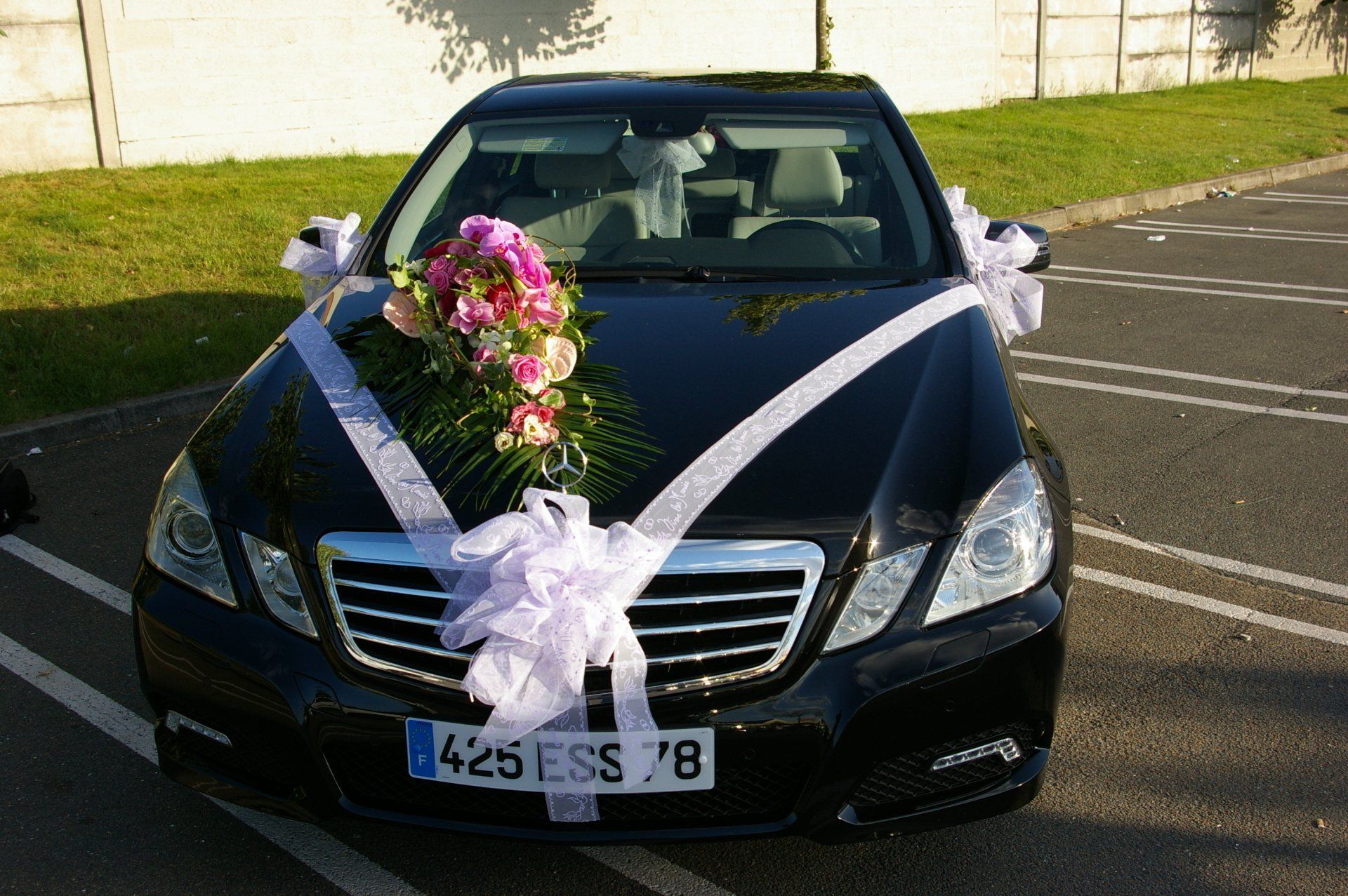 Voiture noire décorée de rubans blancs et de fleurs roses, garée sur un parking.