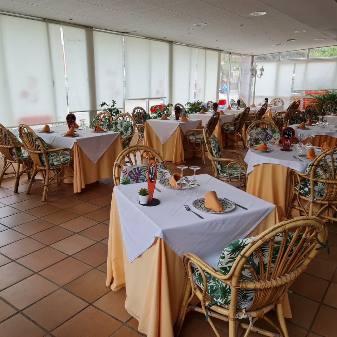 Comedor de restaurante con mesas preparadas para el servicio, sillas de mimbre, mantelería color amarillo claro, sobrecubiertas blancas, suelo de baldosas.