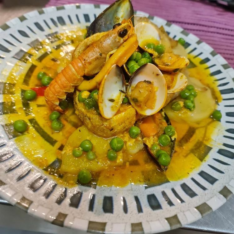 Guiso de mariscos con camarones, almejas, mejillones, guisantes y patatas en un caldo amarillo sobre un plato estampado.